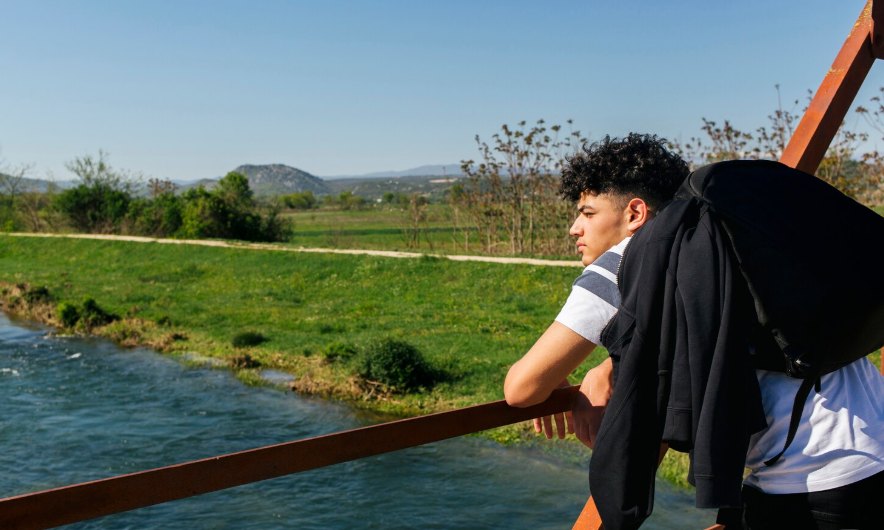 A teenager leaning on a rusty rail beside a river and green meadow